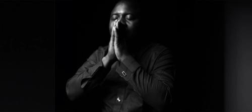 CC- A black and white image of a man in a dress shirt with his eyes closed and his hands in prayer.