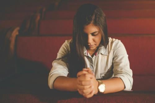 CC- A young woman sits in a church pew alone with her eyes closed and her hands clasped in prayer.