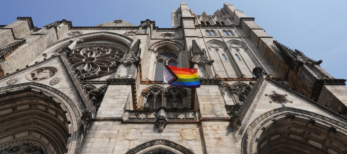 CC-  Looking up the face of a stone gothic-style church with intricate designs and many windows and outcroppings and boasting a modern all-inclusive flag (LGBTQA+)