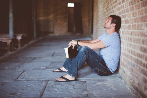 CC- A man sits on the floor against a brick wall with his eyes closed, holding a book in his hands.