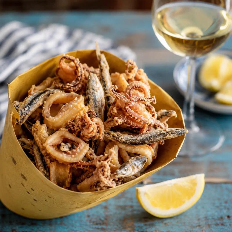 Un cartoccio a cono pieno di pesce fritto misto dorato e croccante, tenuto in mano con il mare di Riomaggiore sullo sfondo