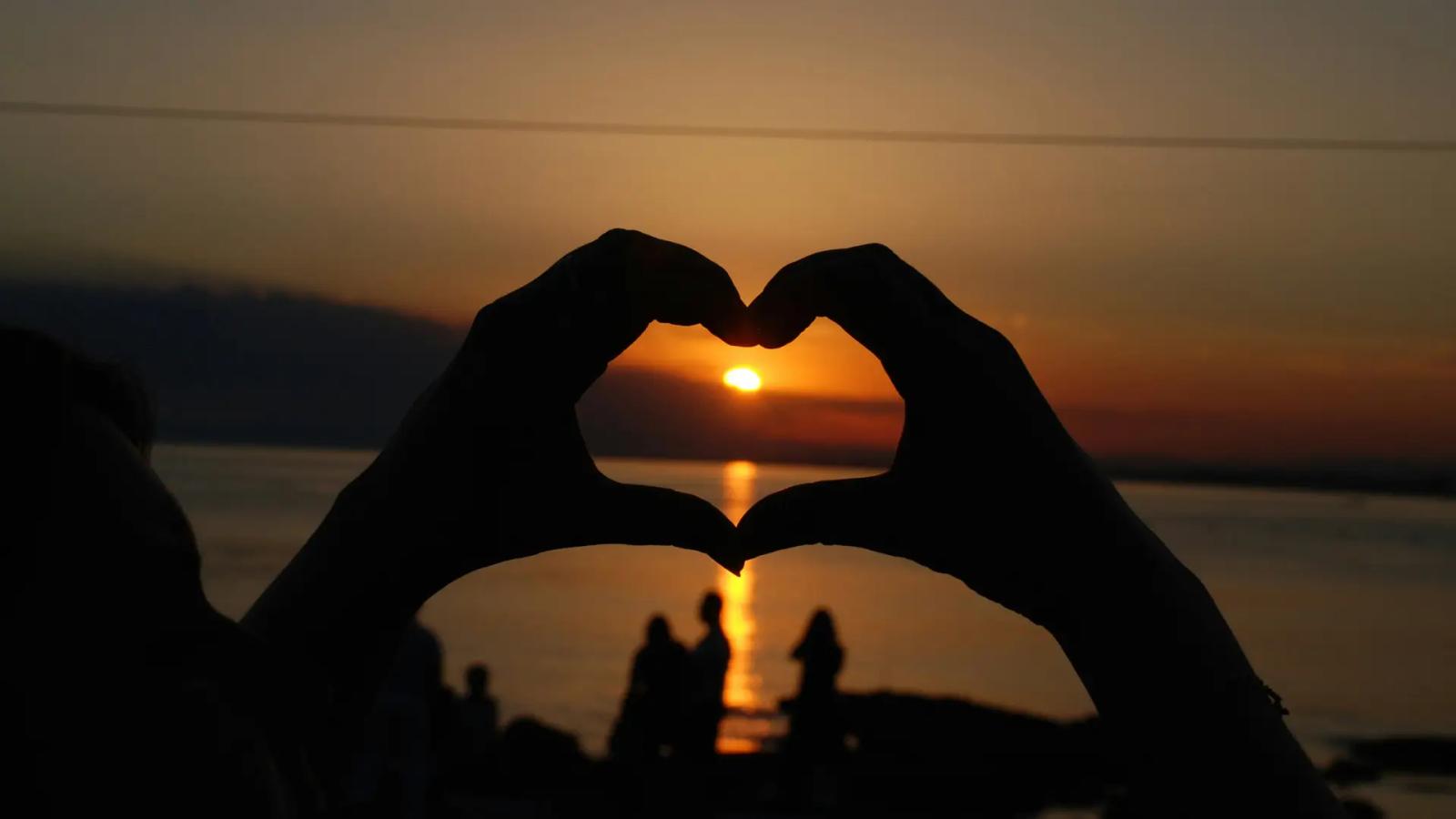 Due mani che formano un cuore davanti a un tramonto sulle Cinque Terre, con il mare e il cielo illuminati da calde sfumature dorate.