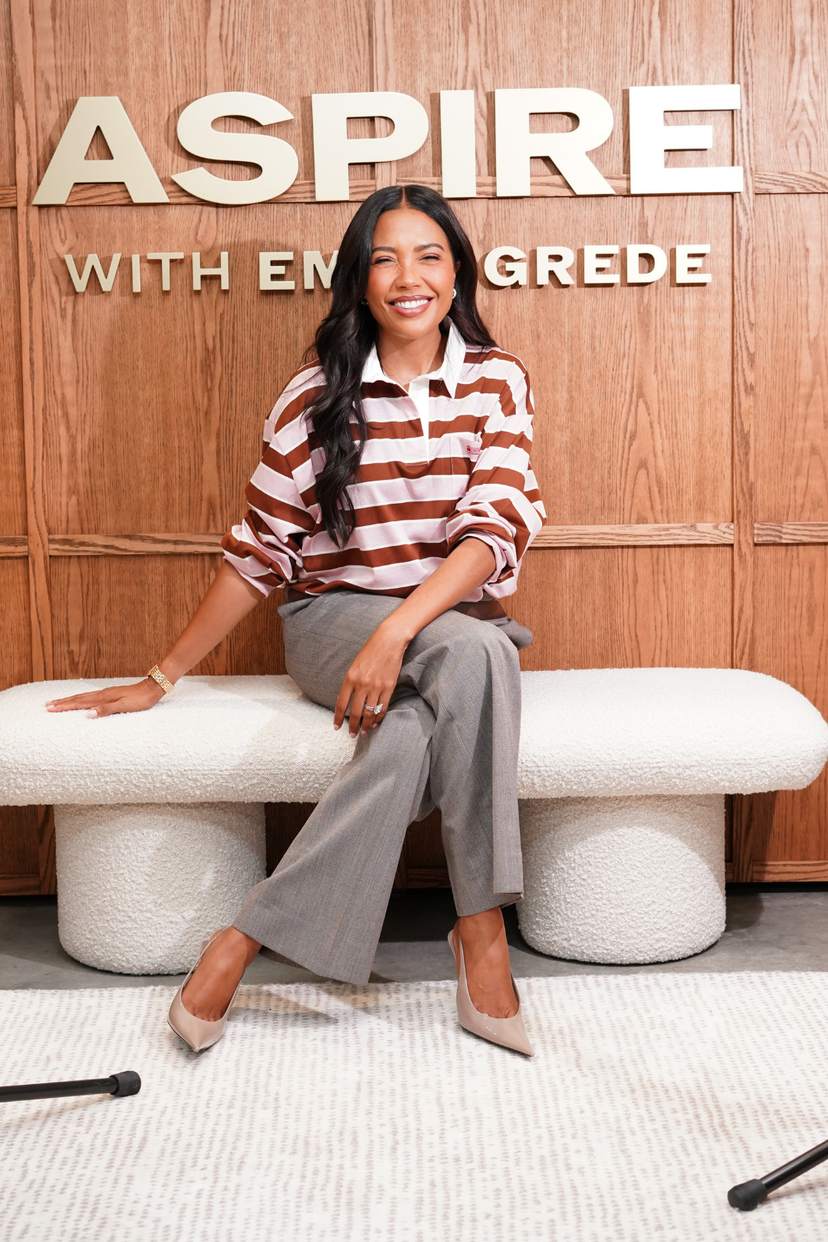 A smiling woman in a striped shirt and gray pants sits on a white bench in front of a wooden wall with "ASPIRE WITH EM GREDE" written on it.