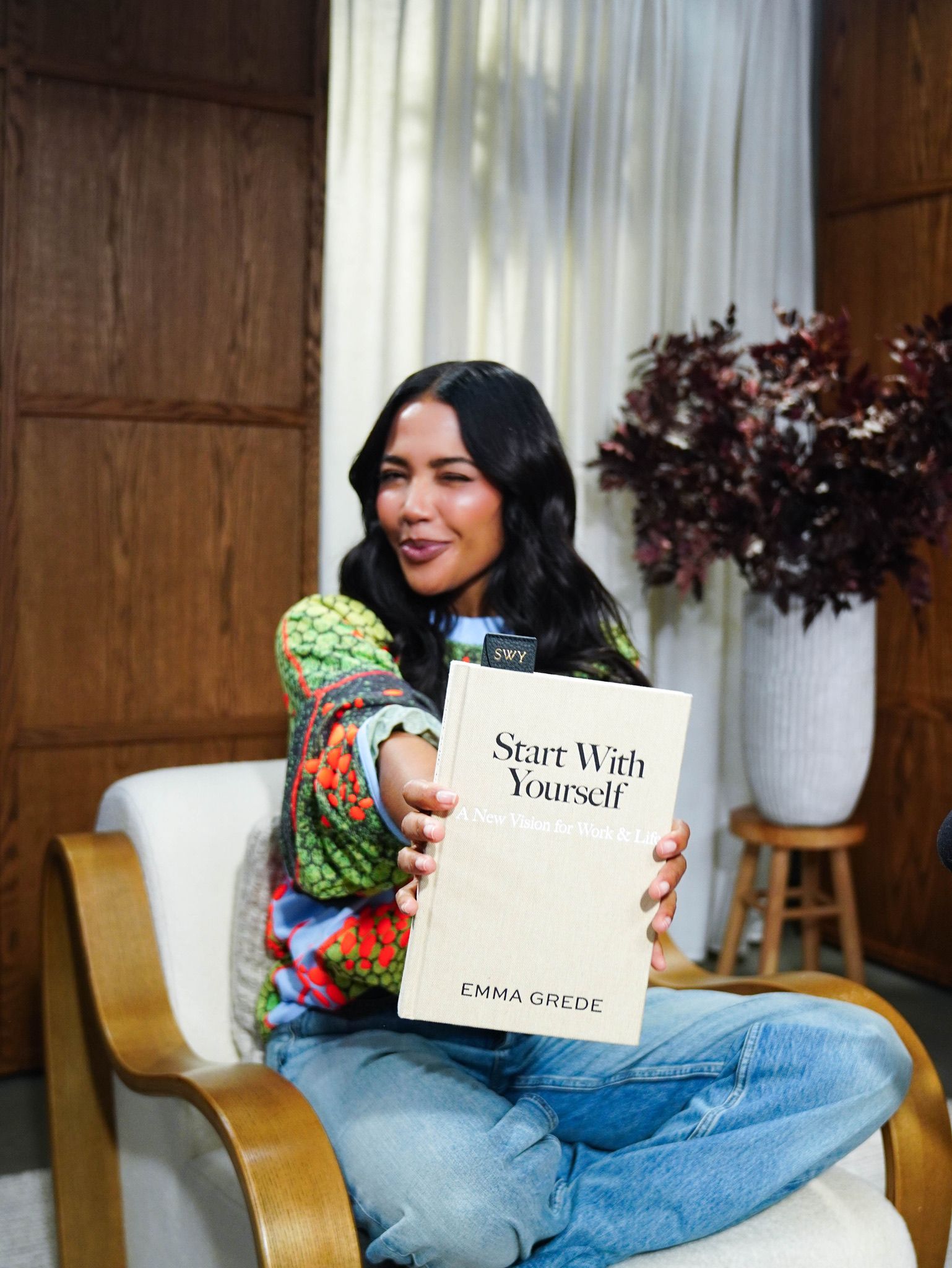 Emma Grede sitting, winking, and holding out her book "Start With Yourself".