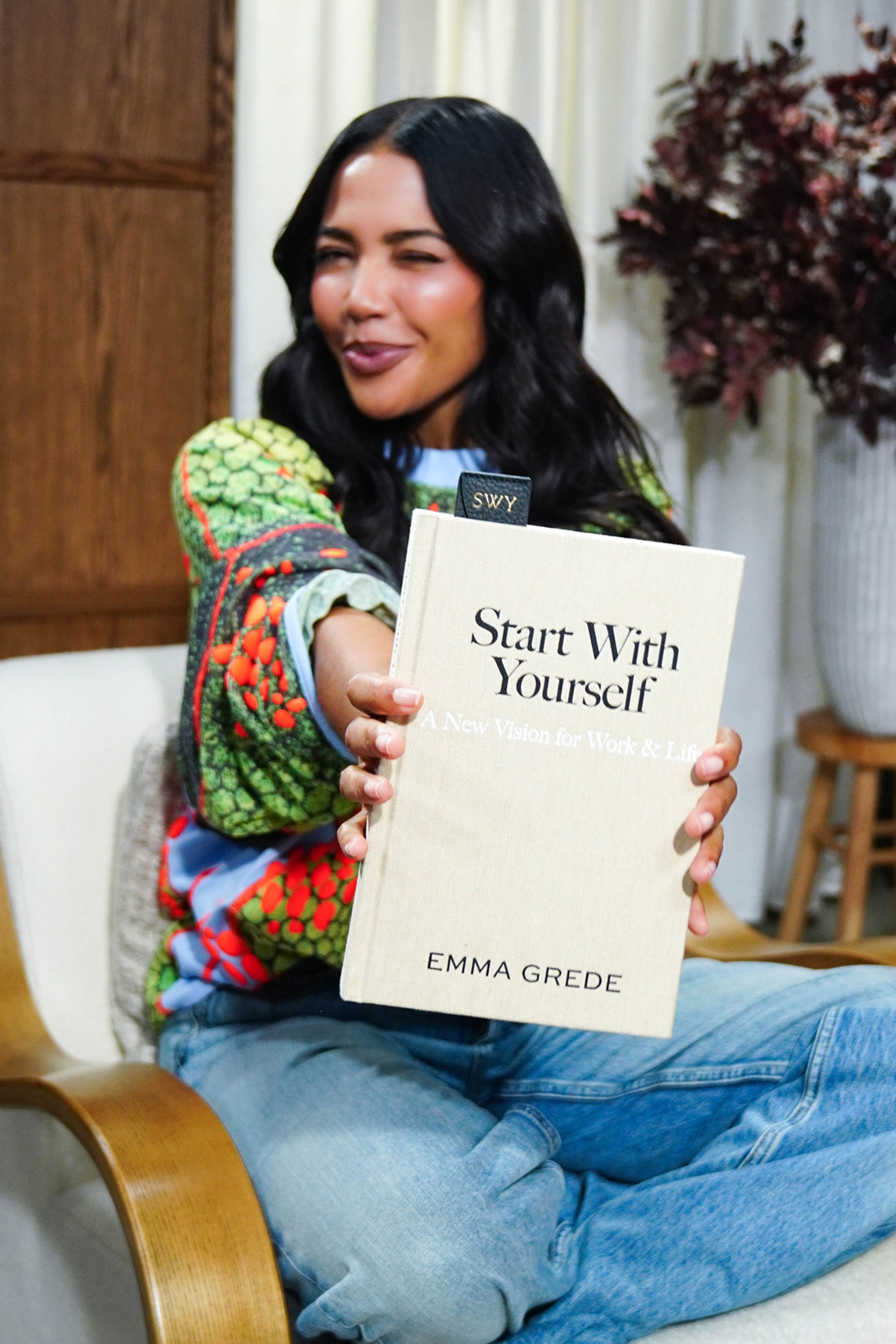 Emma Grede sitting, winking, and holding out her book "Start With Yourself".