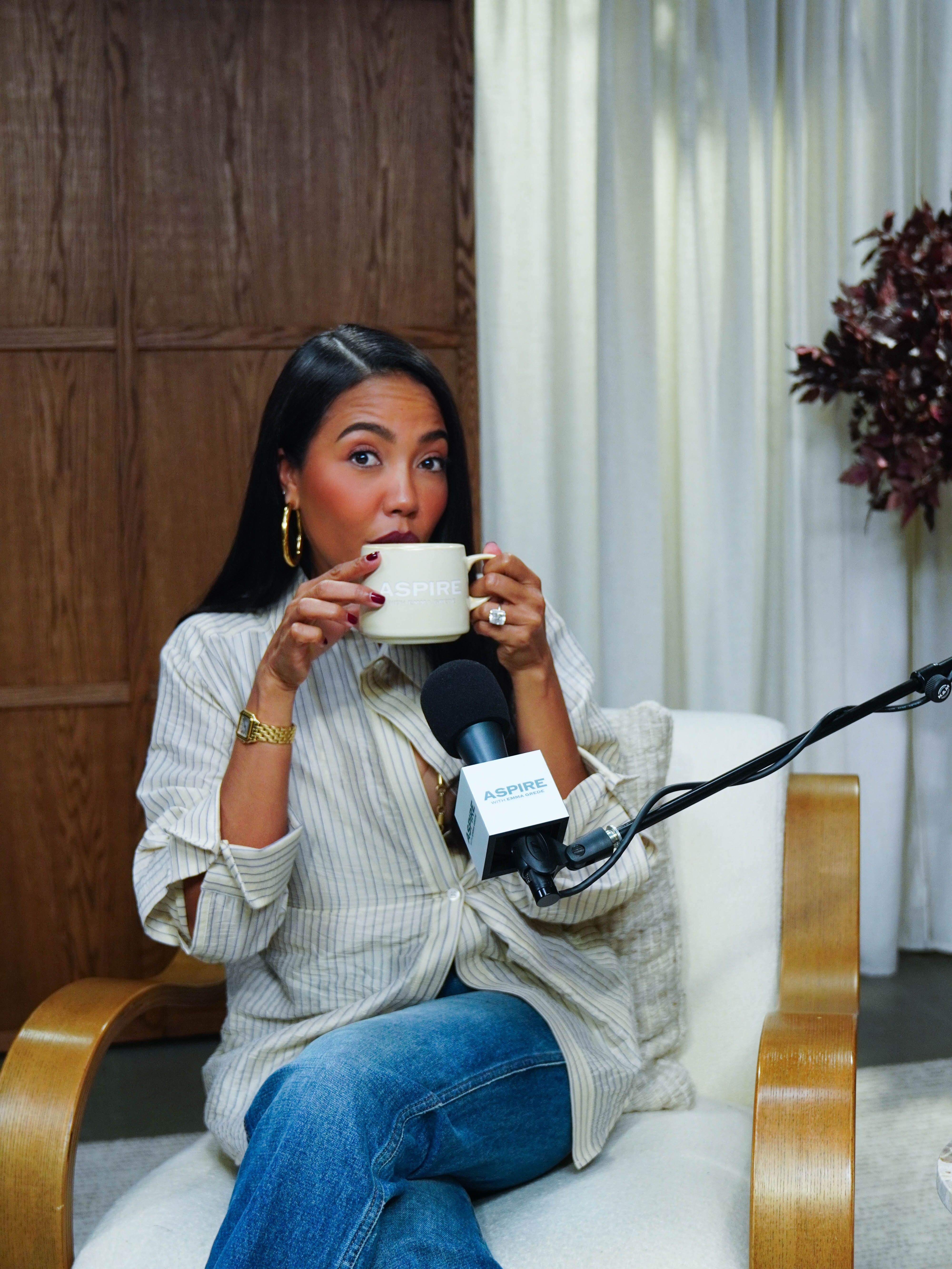 A woman in a striped shirt and jeans sips from an "ASPIRE" mug while seated, with an "ASPIRE" microphone in front of her.