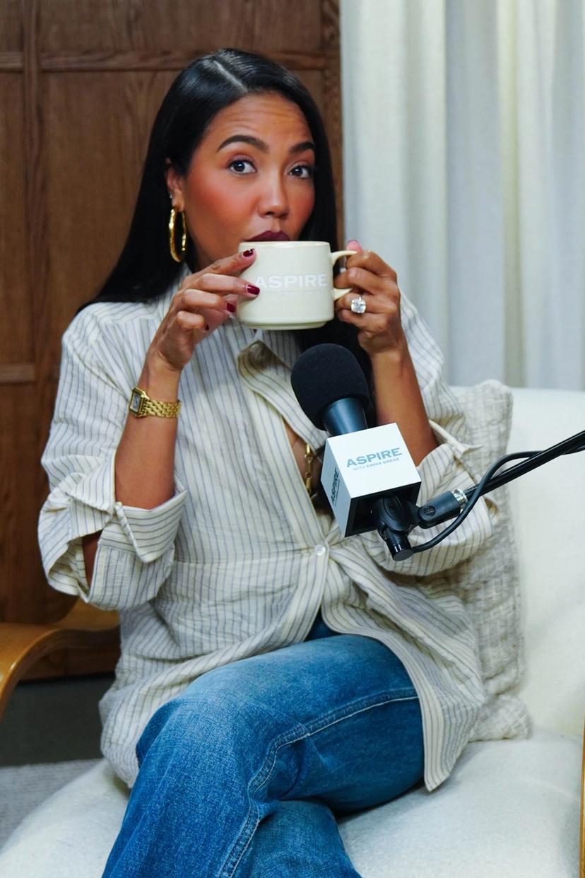 A woman in a striped shirt and jeans sips from an "ASPIRE" mug while seated, with an "ASPIRE" microphone in front of her.