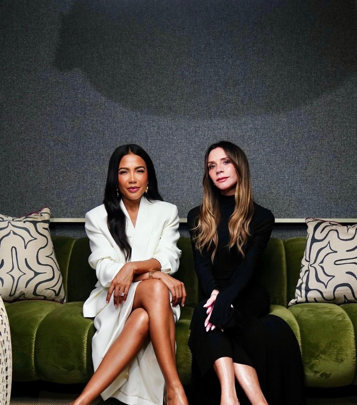 Victoria Beckham in black and a woman with long dark hair in a white blazer sit on a green couch.