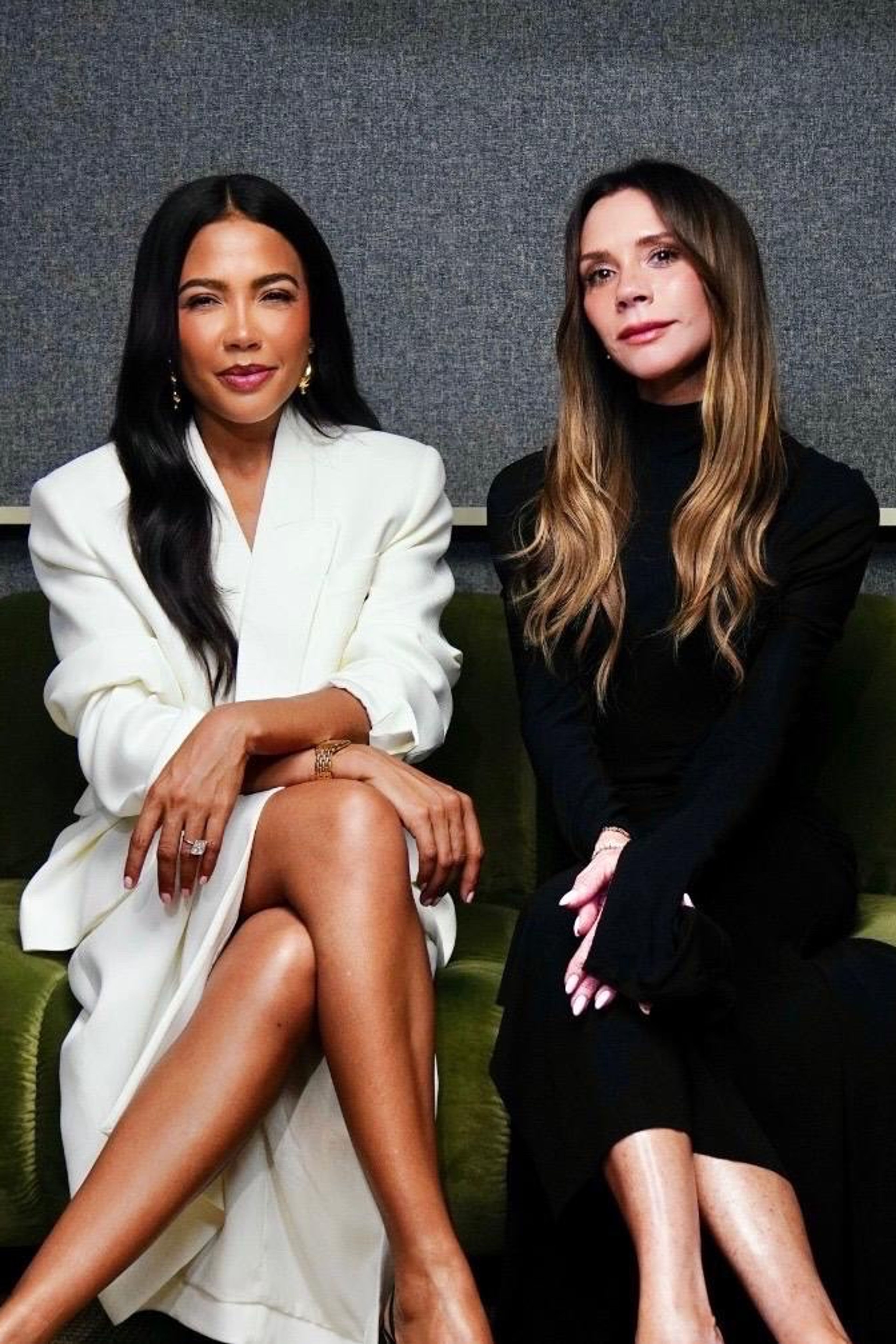 Victoria Beckham in black and a woman with long dark hair in a white blazer sit on a green couch.