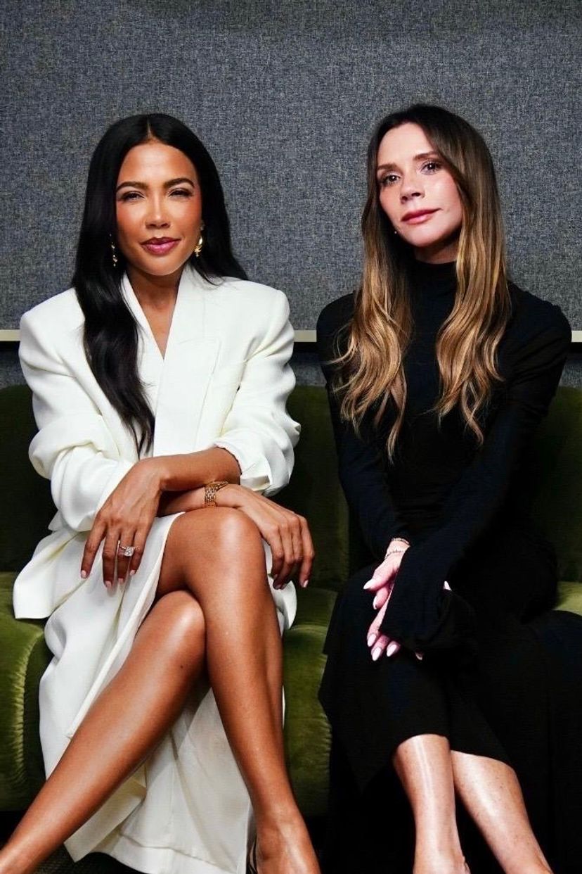 Victoria Beckham in black and a woman with long dark hair in a white blazer sit on a green couch.
