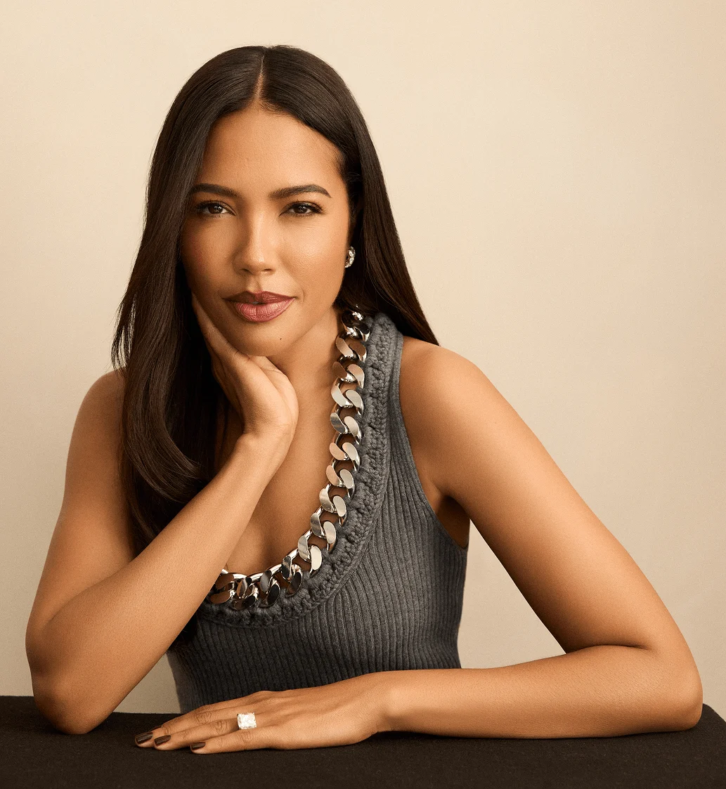A woman with long dark hair, wearing a gray ribbed tank top and a chunky silver chain necklace, rests her chin on her hand, displaying a large ring.