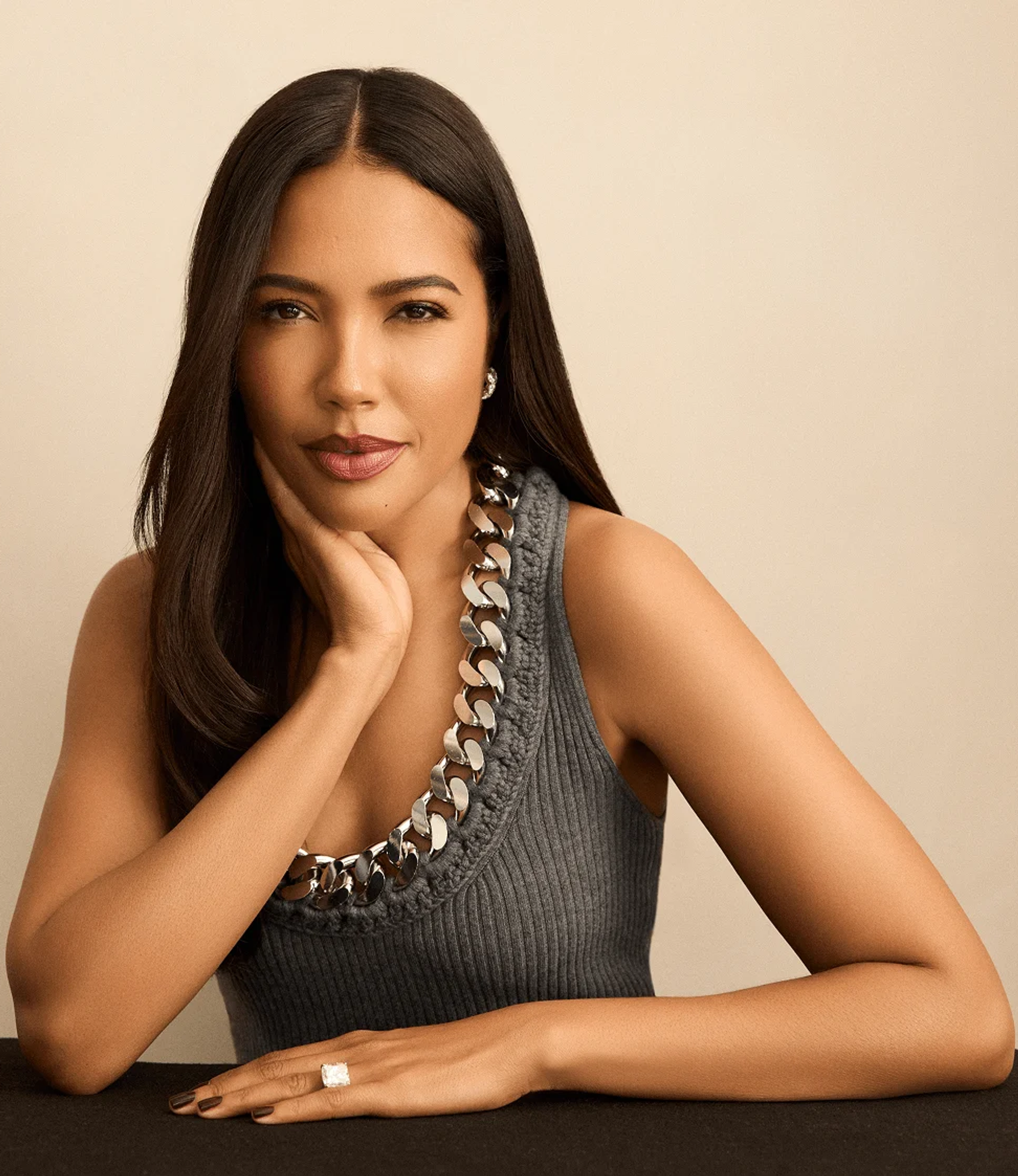 A woman with long dark hair, wearing a gray ribbed tank top and a chunky silver chain necklace, rests her chin on her hand, displaying a large ring.