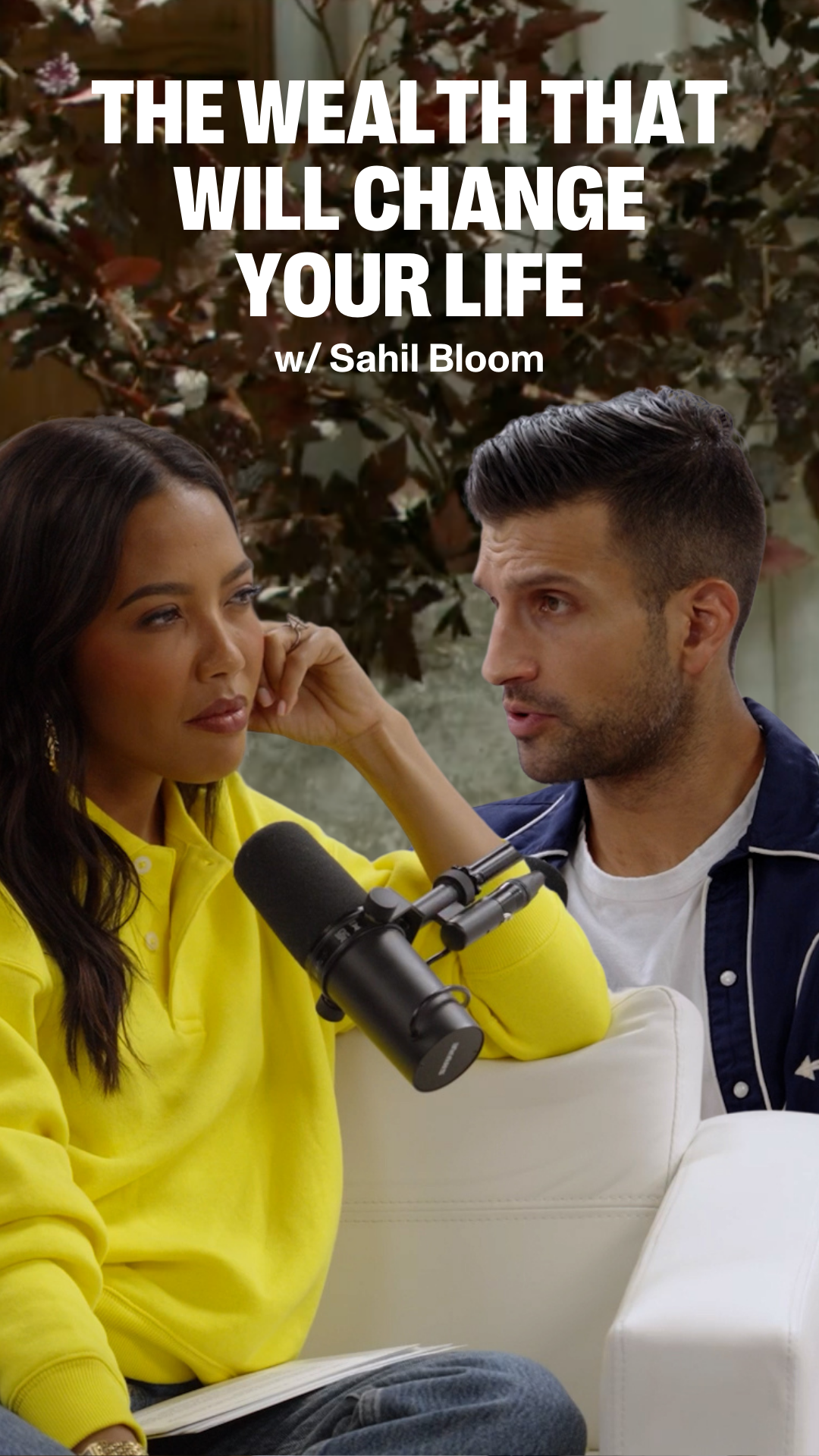 Emma Grede in a yellow top listens to a man speaking, with a microphone between them. Overlay text reads "THE WEALTH THAT WILL CHANGE YOUR LIFE w/ Sahil Bloom".
