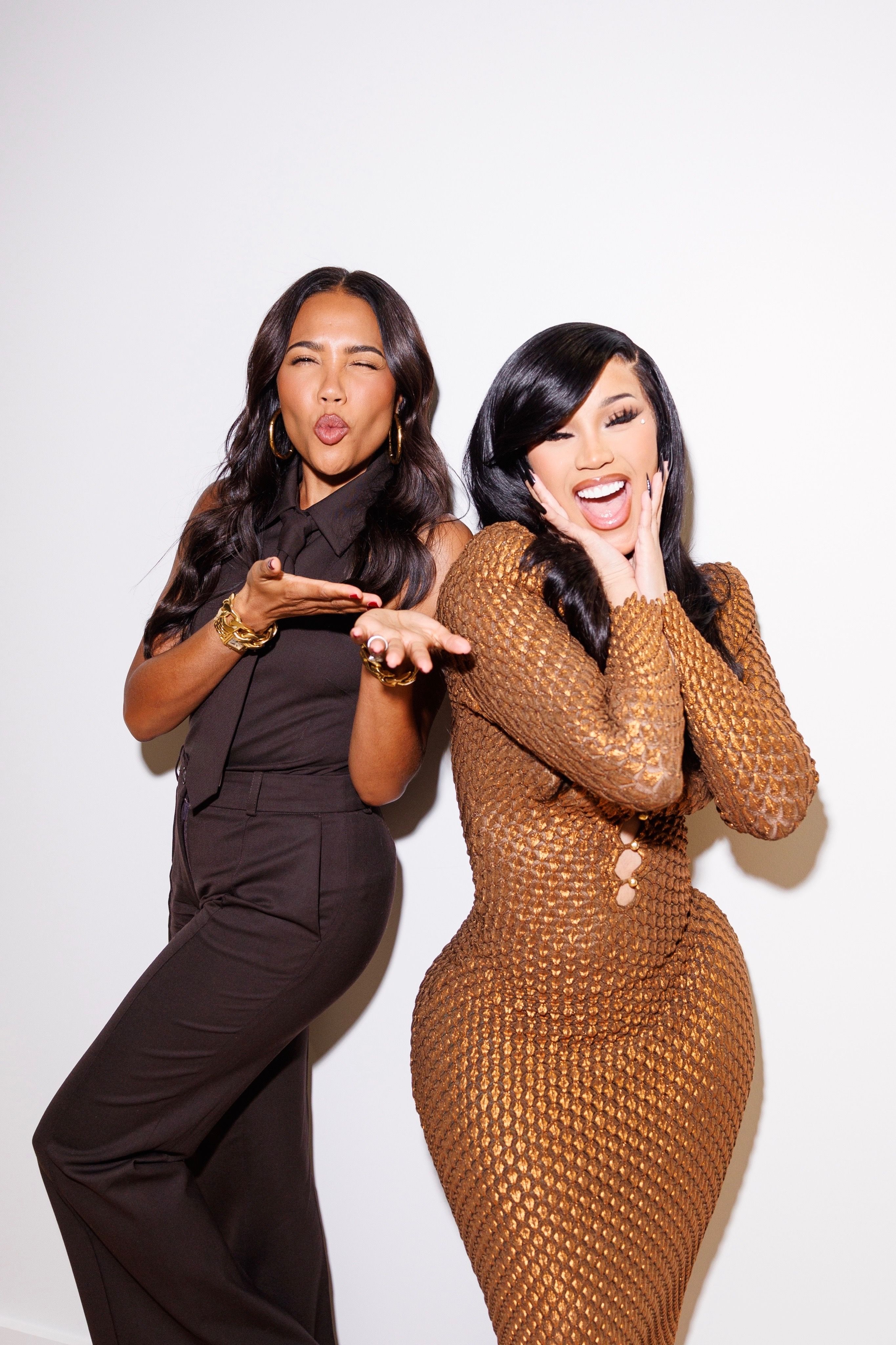 Two women playfully posing against a white background. The woman on the left, emma grede, in a dark brown outfit, makes a kiss face while the woman on the right, cardi b, in a bronze textured dress, has a wide open-mouthed smile with hands on her cheeks.