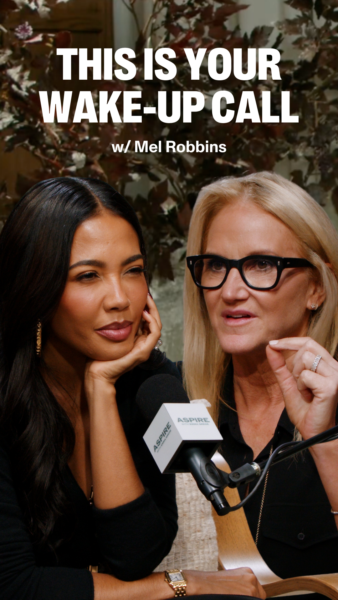 Two women record a podcast; Mel Robbins, a blonde woman in glasses, speaks and gestures next to a dark-haired woman, Emma Grede listening. Text reads "THIS IS YOUR WAKE-UP CALL w/ Mel Robbins."