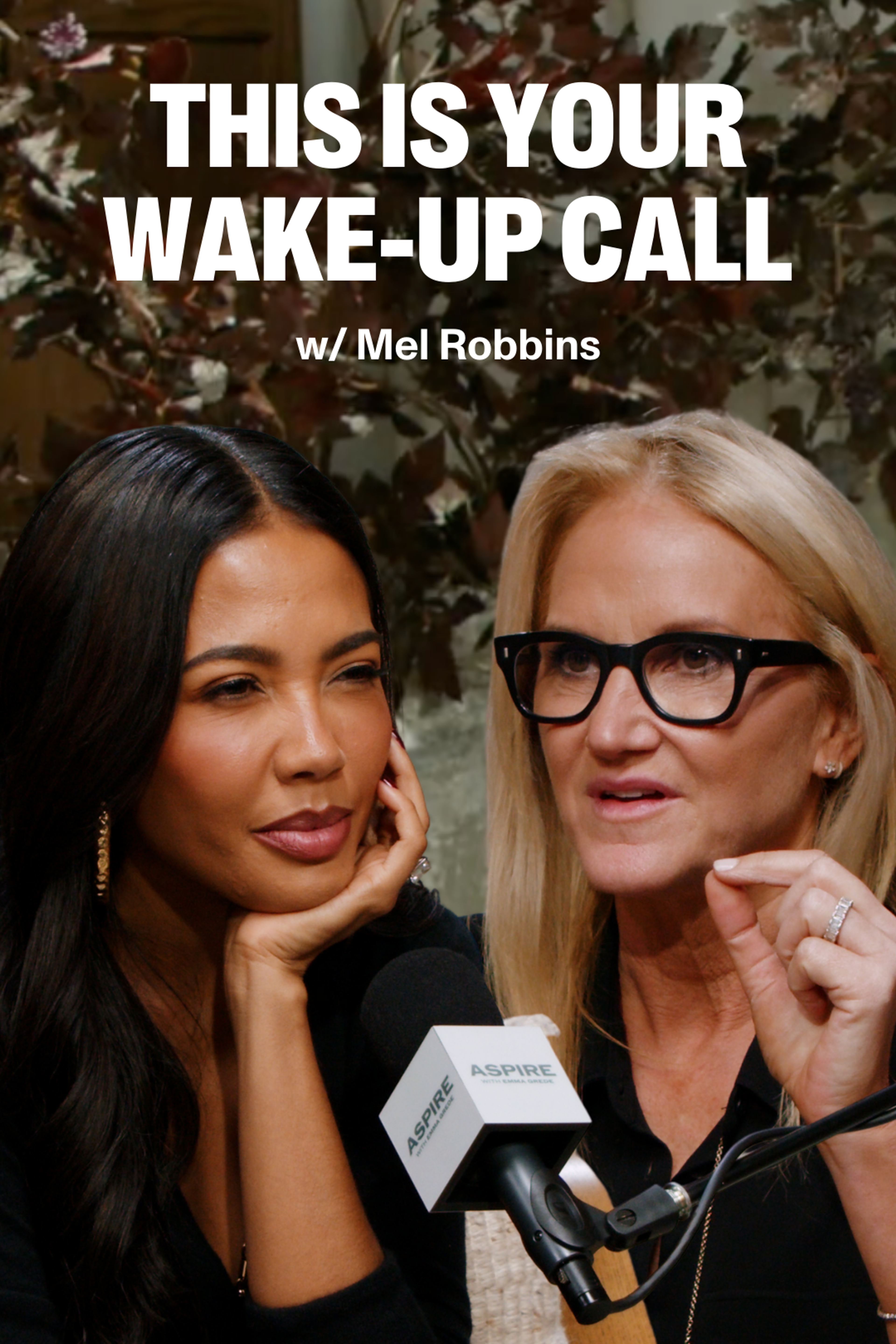 Two women record a podcast; Mel Robbins, a blonde woman in glasses, speaks and gestures next to a dark-haired woman, Emma Grede listening. Text reads "THIS IS YOUR WAKE-UP CALL w/ Mel Robbins."