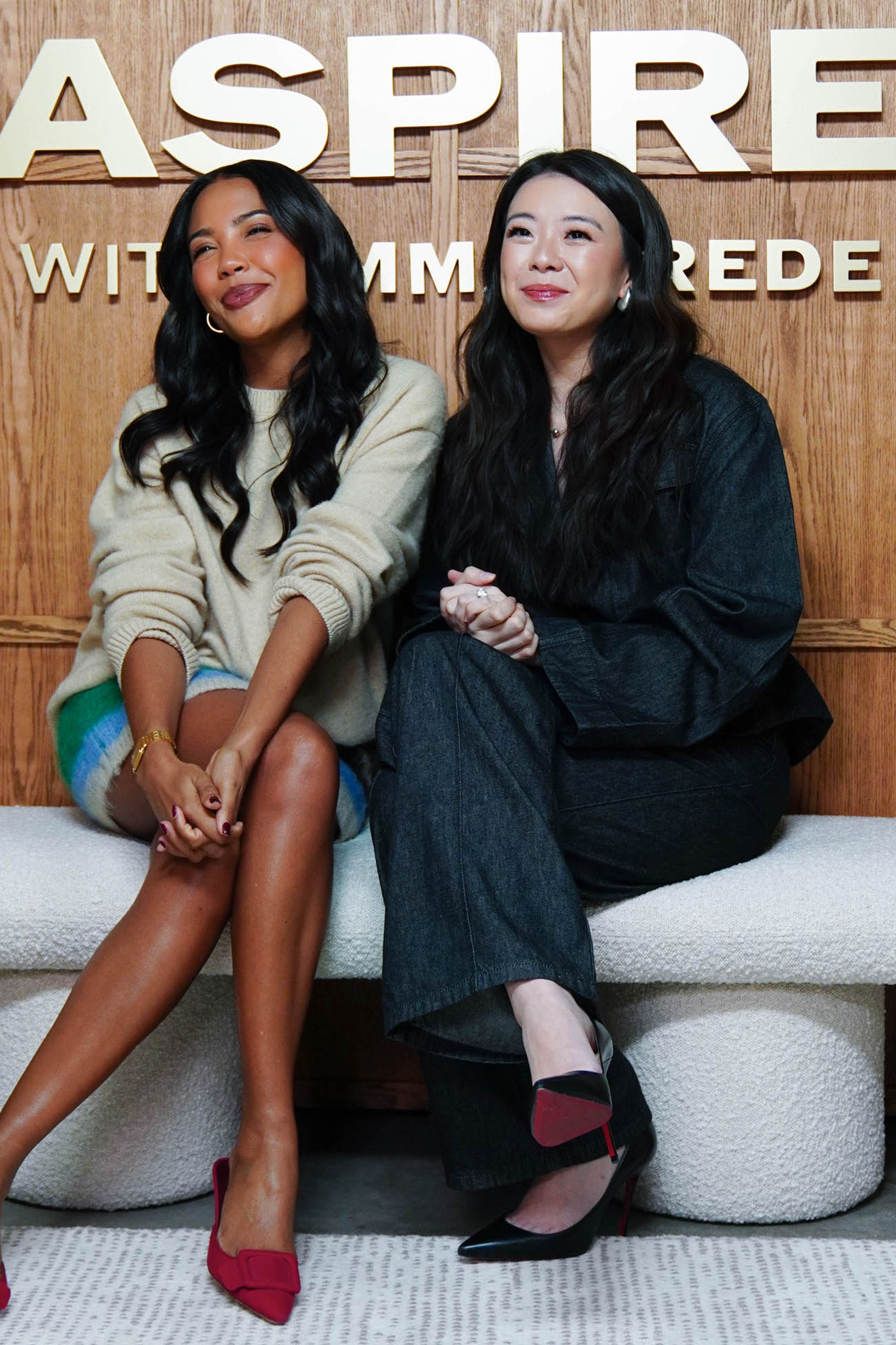 Two women emma grede vivian tu sit side-by-side on a white bench in front of a wooden wall with gold "ASPIRE" lettering.