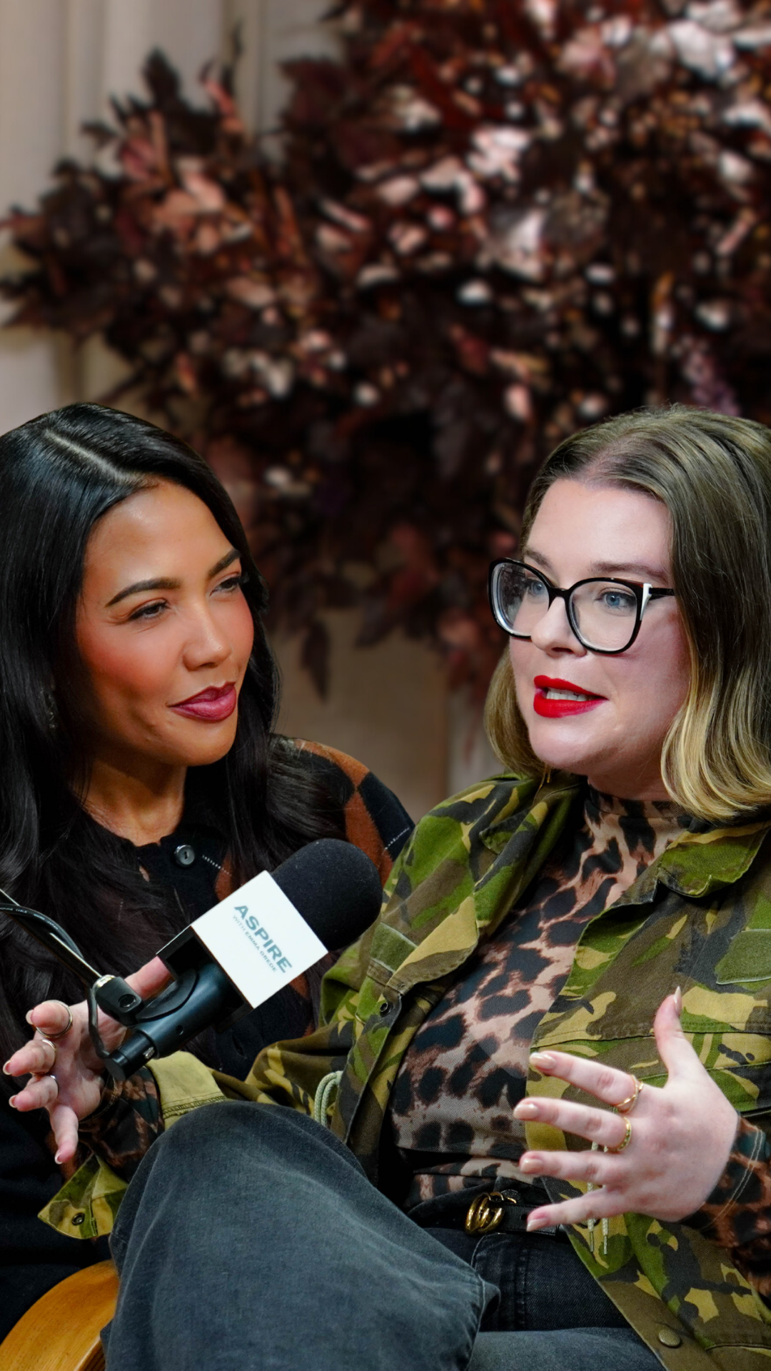A dark-haired woman emma grede smiles at another woman tori dunlap in glasses and red lipstick speaking into a microphone.