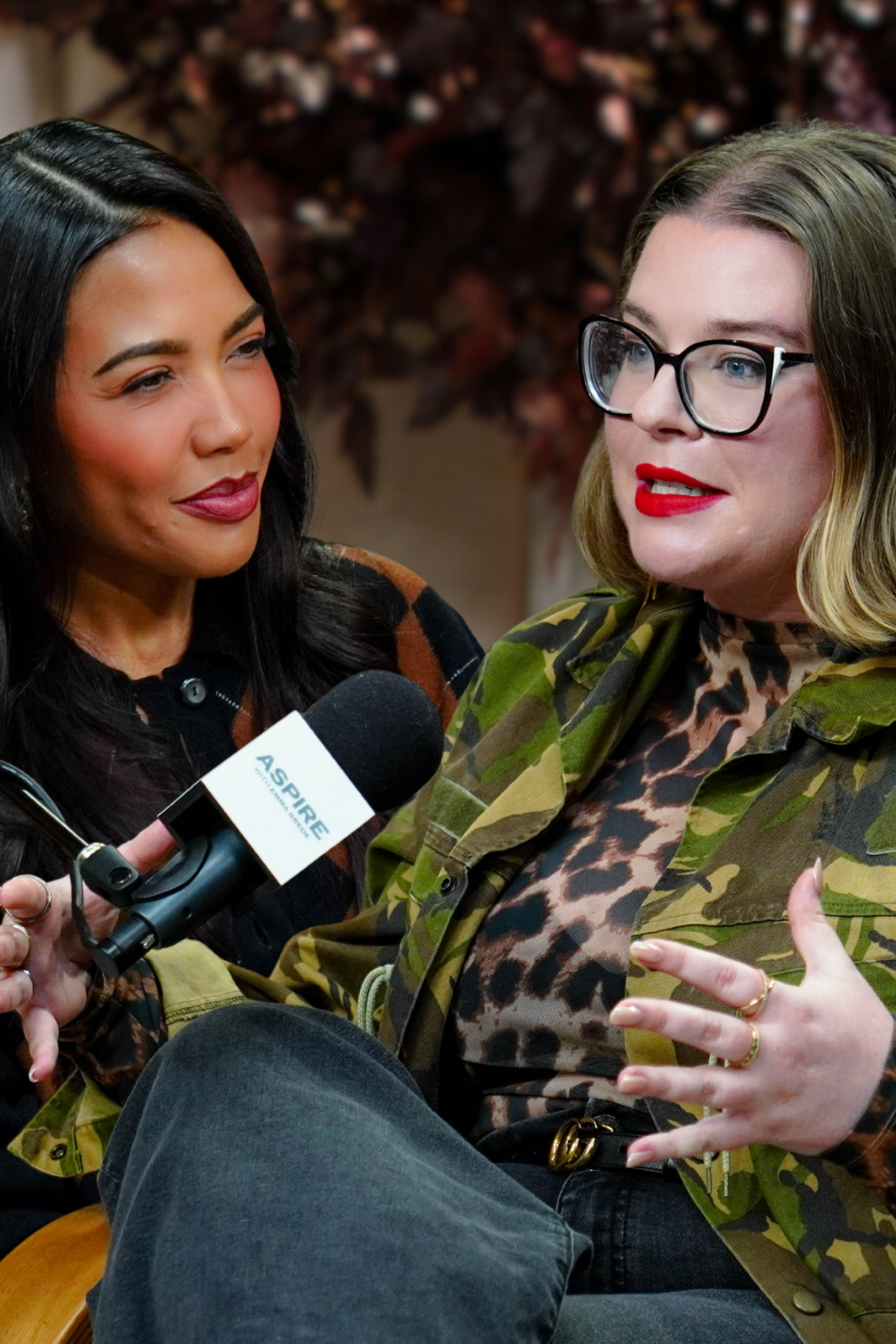 A dark-haired woman emma grede smiles at another woman tori dunlap in glasses and red lipstick speaking into a microphone.