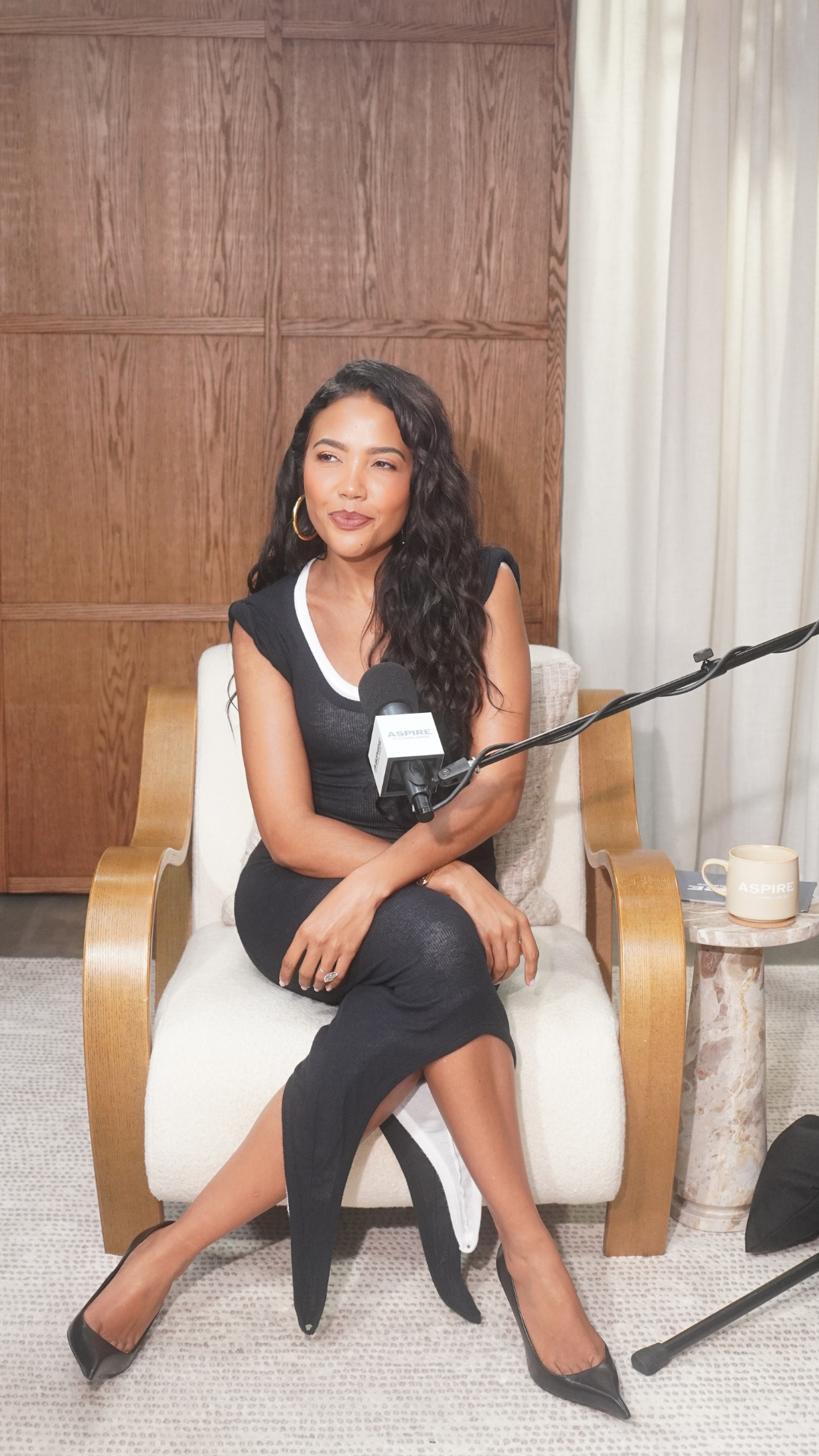 A woman with long wavy hair sits in a chair, holding an ASPIRE microphone, wearing a black dress with white trim and black heels.
