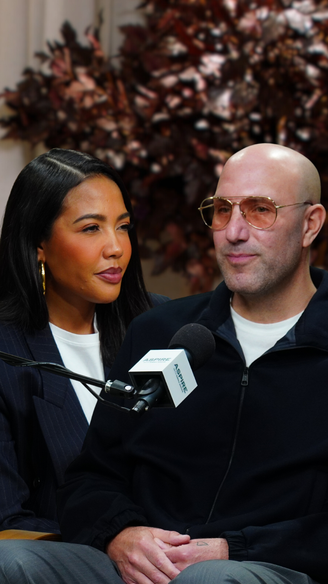 A woman Emma Grede with long dark hair looks towards a bald man Rich Kleiman in tinted glasses, with an ASPIRE microphone between them.