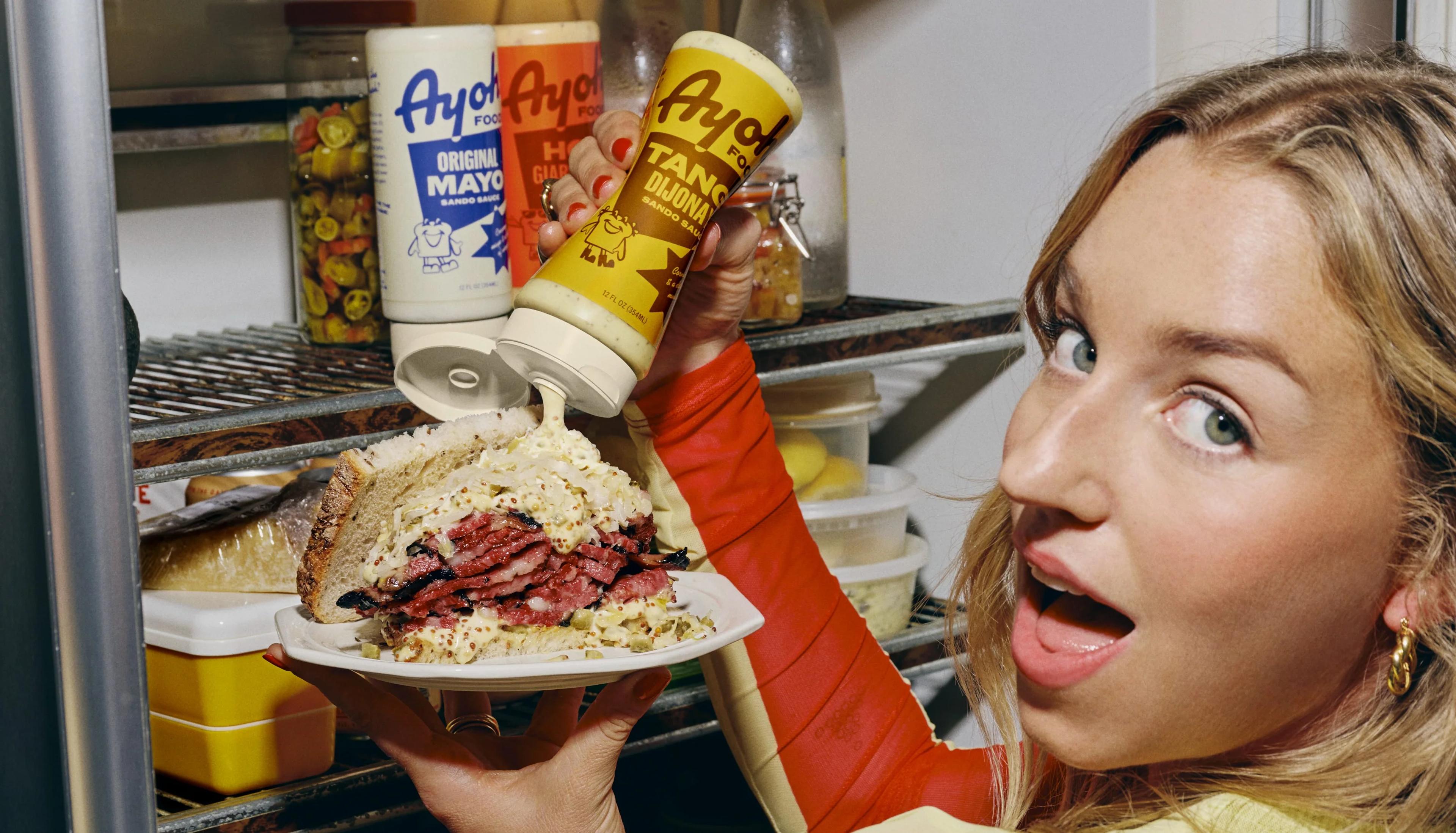 Woman squeezing Ayoh Tangy Dijonnayo sauce onto an overstuffed deli sandwich in a fridge stocked with Ayoh bottles