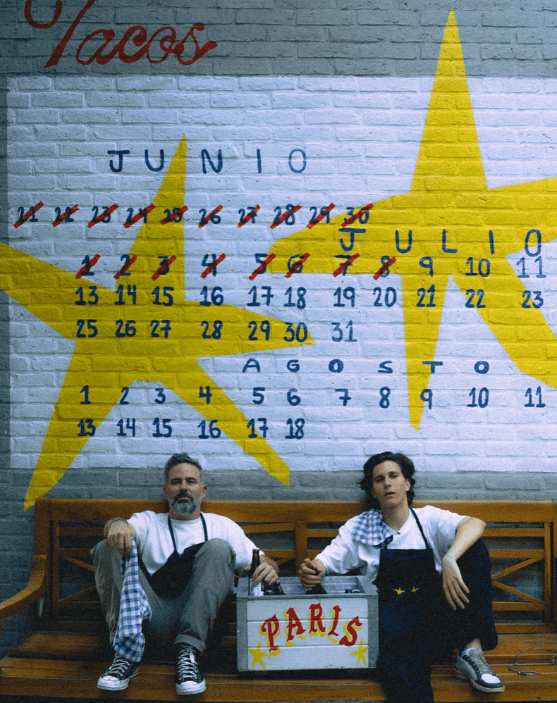 Two apron-wearing founders sitting on a bench beneath a hand-painted calendar wall for taco restaurant Paris