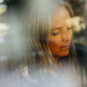Soft-focus portrait of a person with long, graying hair looking down thoughtfully, seen through a hazy, light-filled foreground.
