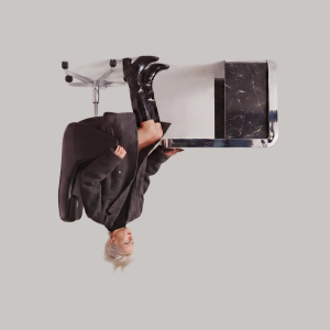 Fashion-forward person with short platinum hair reclines in a modern office chair, wearing an oversized dark coat and shiny black knee-high boots, next to a sleek black marble and chrome desk against a plain background.