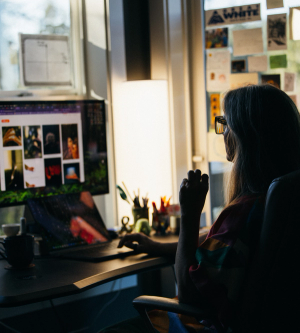Person working at a dimly lit desk, looking at a large computer monitor displaying a grid of colorful images, with notes and photos pinned to the wall behind them.