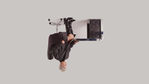 A stylish person in an oversized gray coat and black patent boots lounges in a modern office chair with their feet up on a sleek black marble and chrome desk against a plain light background.