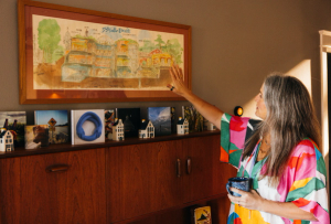 A woman in a colorful dress stands by a wooden cabinet, reaching up to gesture toward a large framed illustration on the wall while holding a blue mug in her other hand.