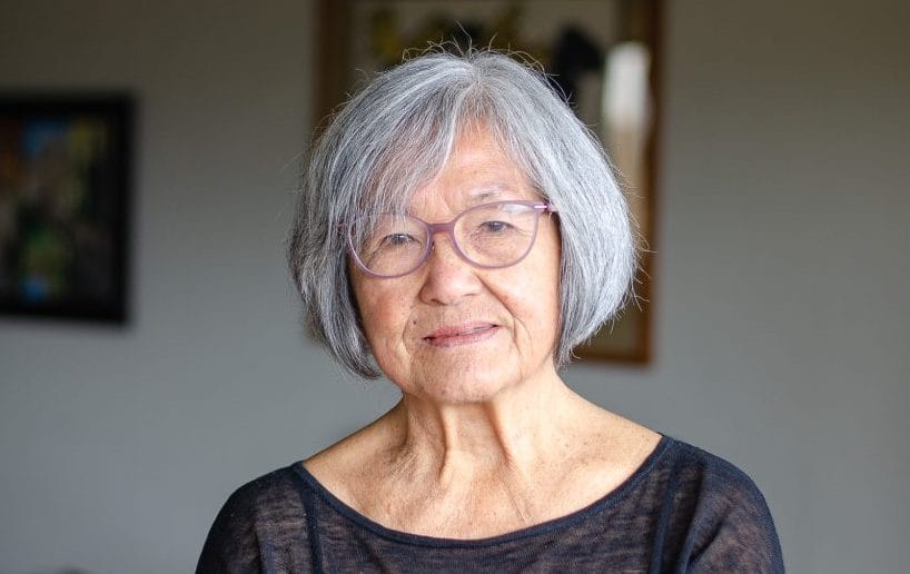 Nellie Wong with a slight smile. She is an older person wearing a dark shirt. She wears translucent lavender glasses and her grey hair is in a medium bob.