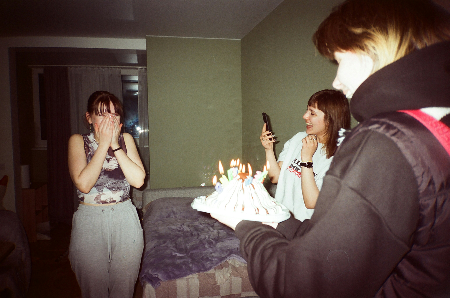 Three young women celebrating a birthday with a lit cake; one is surprised, another takes a photo.