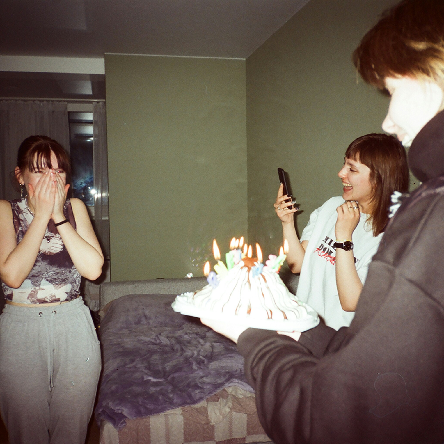 Three young women celebrating a birthday with a lit cake; one is surprised, another takes a photo.