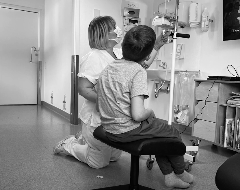 En maskerad sjuksköterska knäböjer bredvid ett sittande barn, båda fokuserade på ett droppställ.