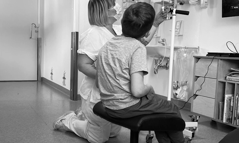 En maskerad sjuksköterska knäböjer bredvid ett sittande barn, båda fokuserade på ett droppställ.