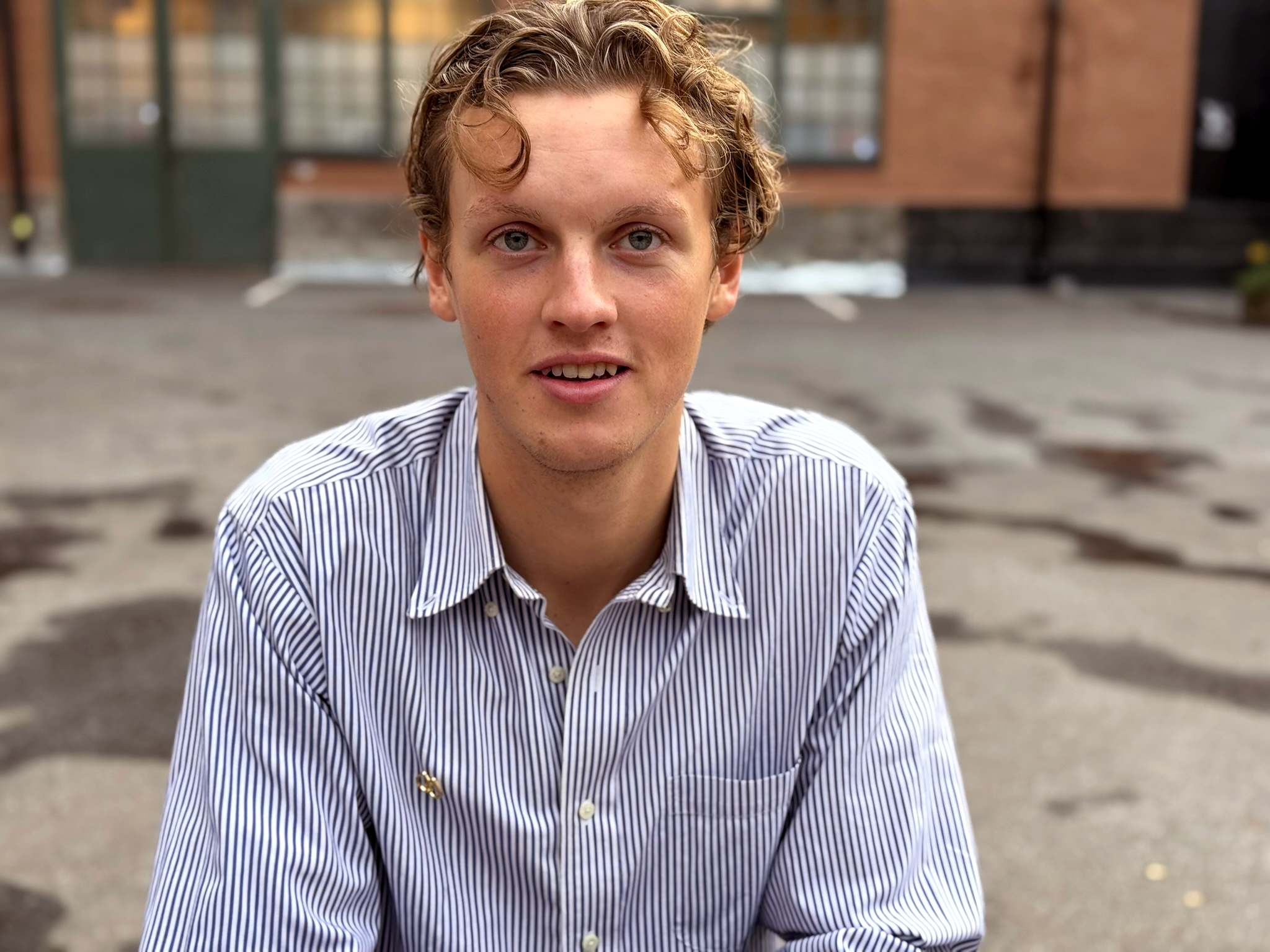 En ung man med lockigt blont hår iklädd en randig skjorta ser in i kameran med ett lätt leende.