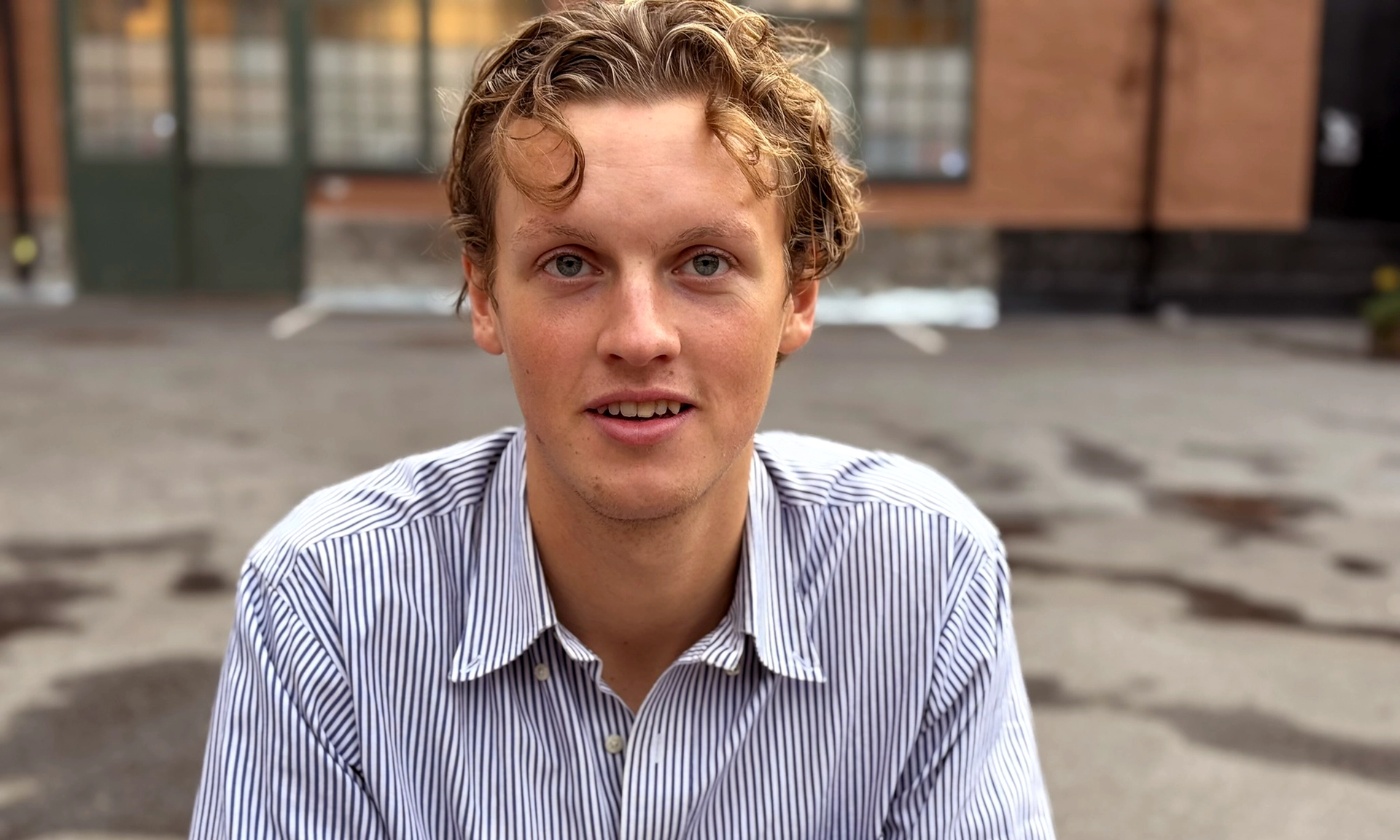 En ung man med lockigt blont hår iklädd en randig skjorta ser in i kameran med ett lätt leende.