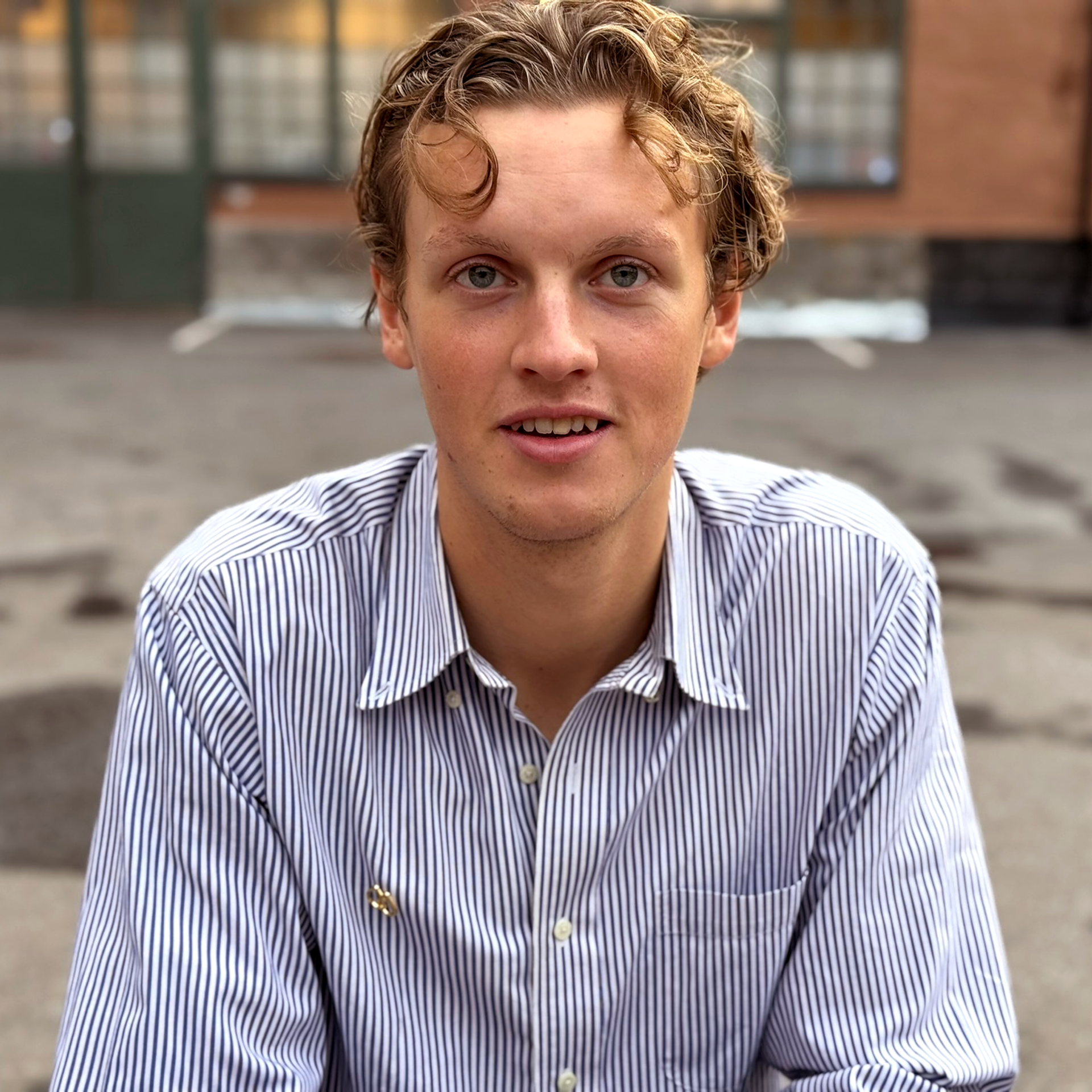 En ung man med lockigt blont hår iklädd en randig skjorta ser in i kameran med ett lätt leende.