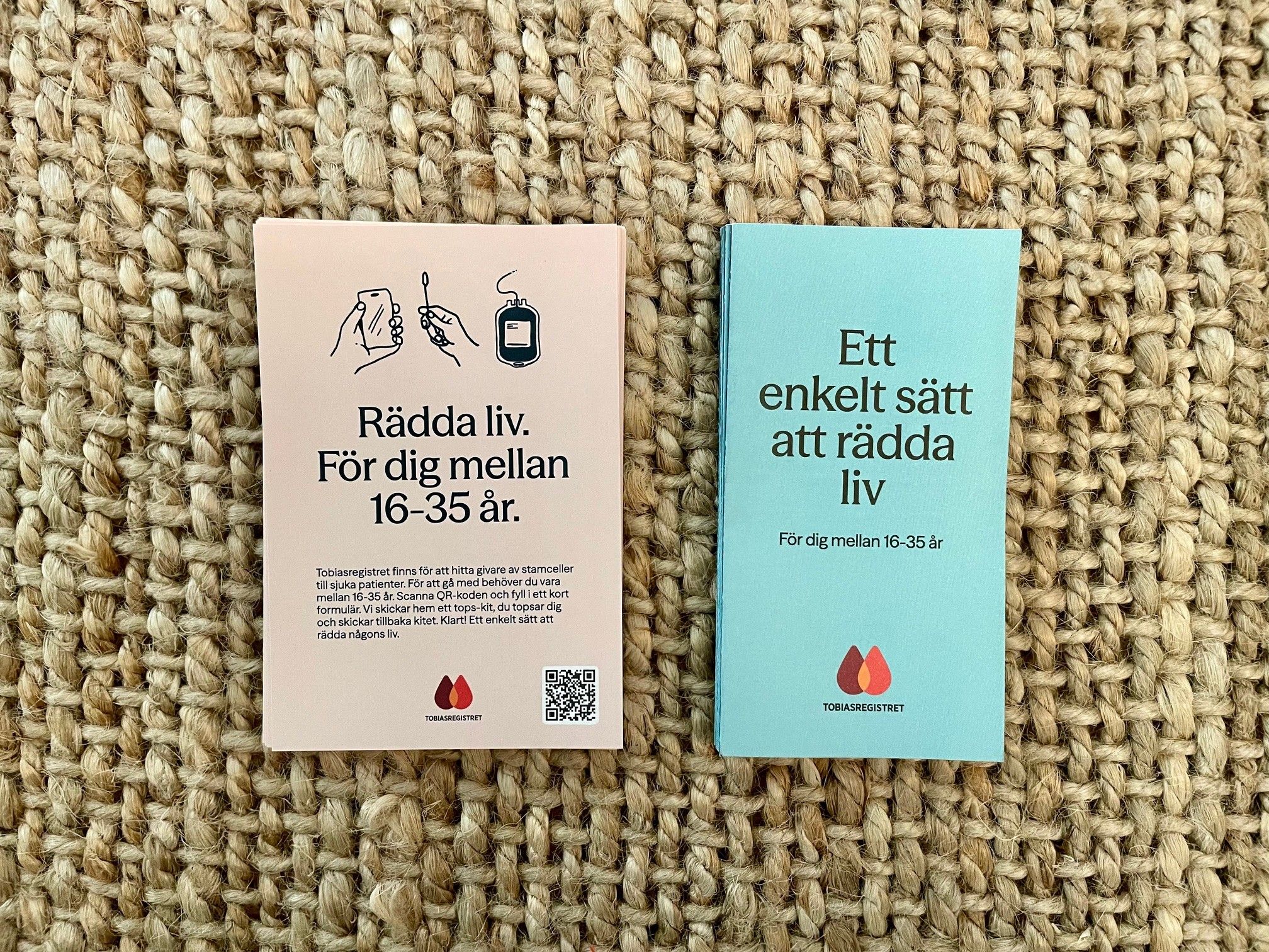 Två svenska broschyrer från Tobiasregistret, en rosa och en blå, som uppmanar personer i åldern 16–35 år att rädda liv genom donation, utlagda på en vävd matta.