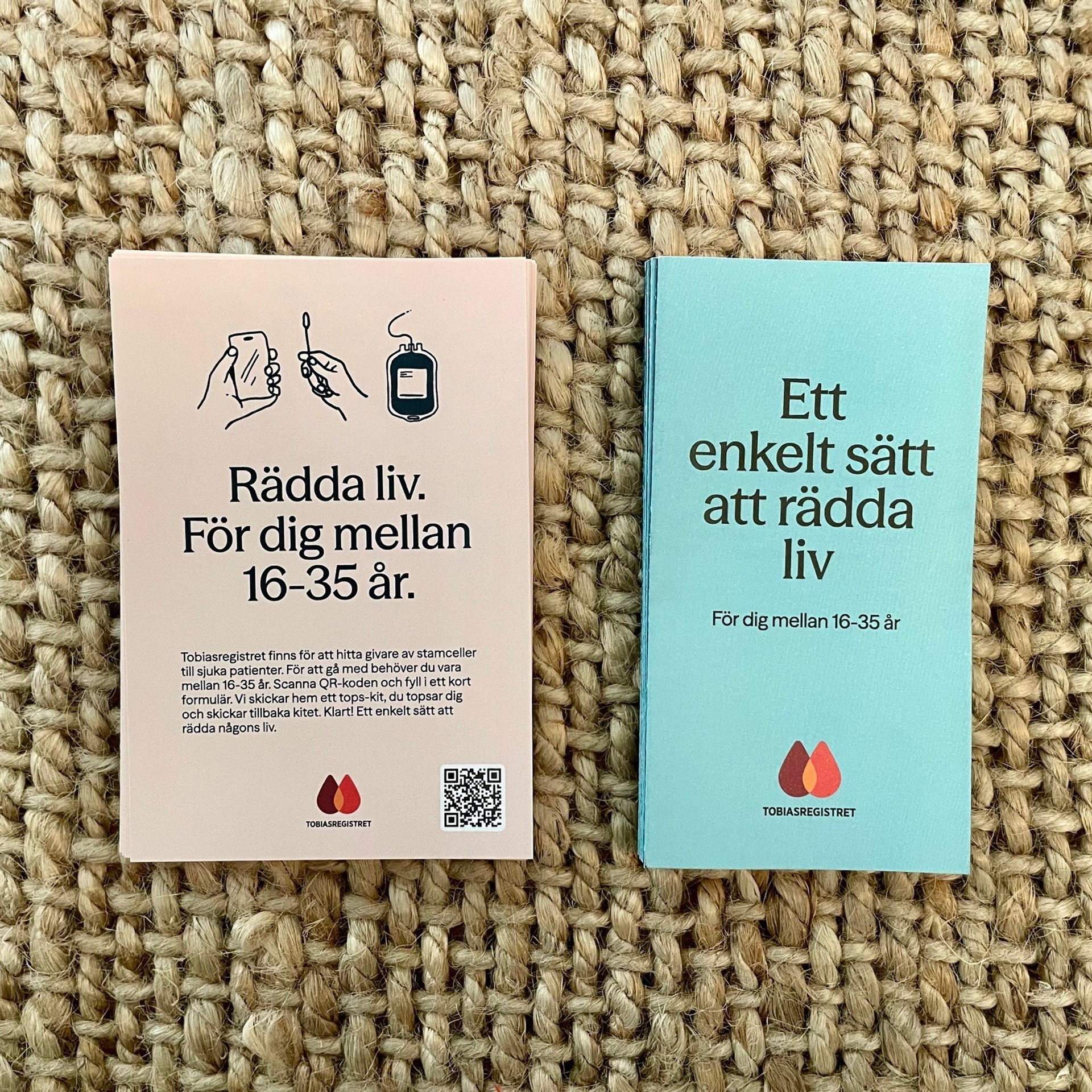 Två svenska broschyrer från Tobiasregistret, en rosa och en blå, som uppmanar personer i åldern 16–35 år att rädda liv genom donation, utlagda på en vävd matta.