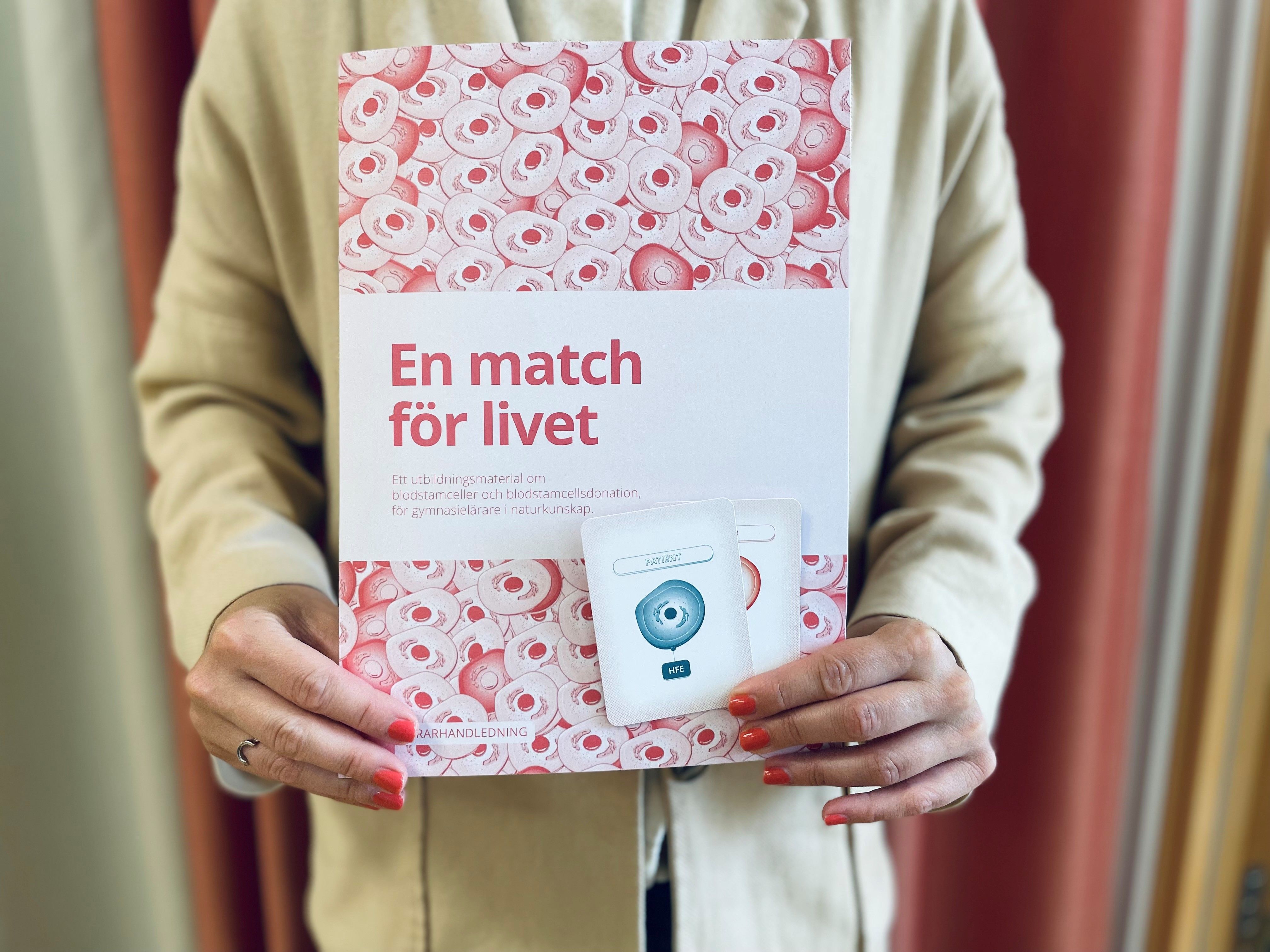 En person håller i ett häfte med ett blodcells-mönster, med titeln ”En match för livet”, och ett vitt paket märkt ”Intyget”.