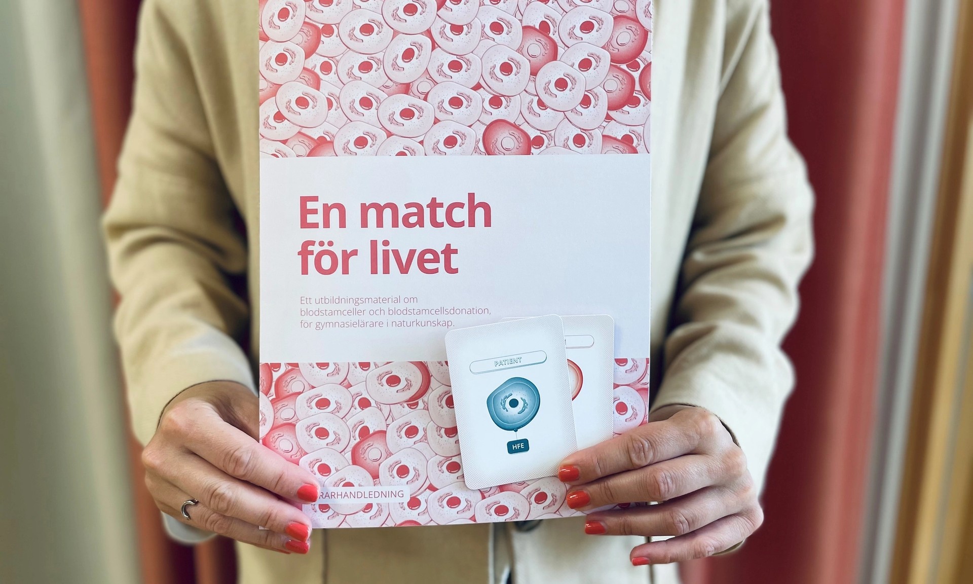 En person håller i ett häfte med ett blodcells-mönster, med titeln ”En match för livet”, och ett vitt paket märkt ”Intyget”.