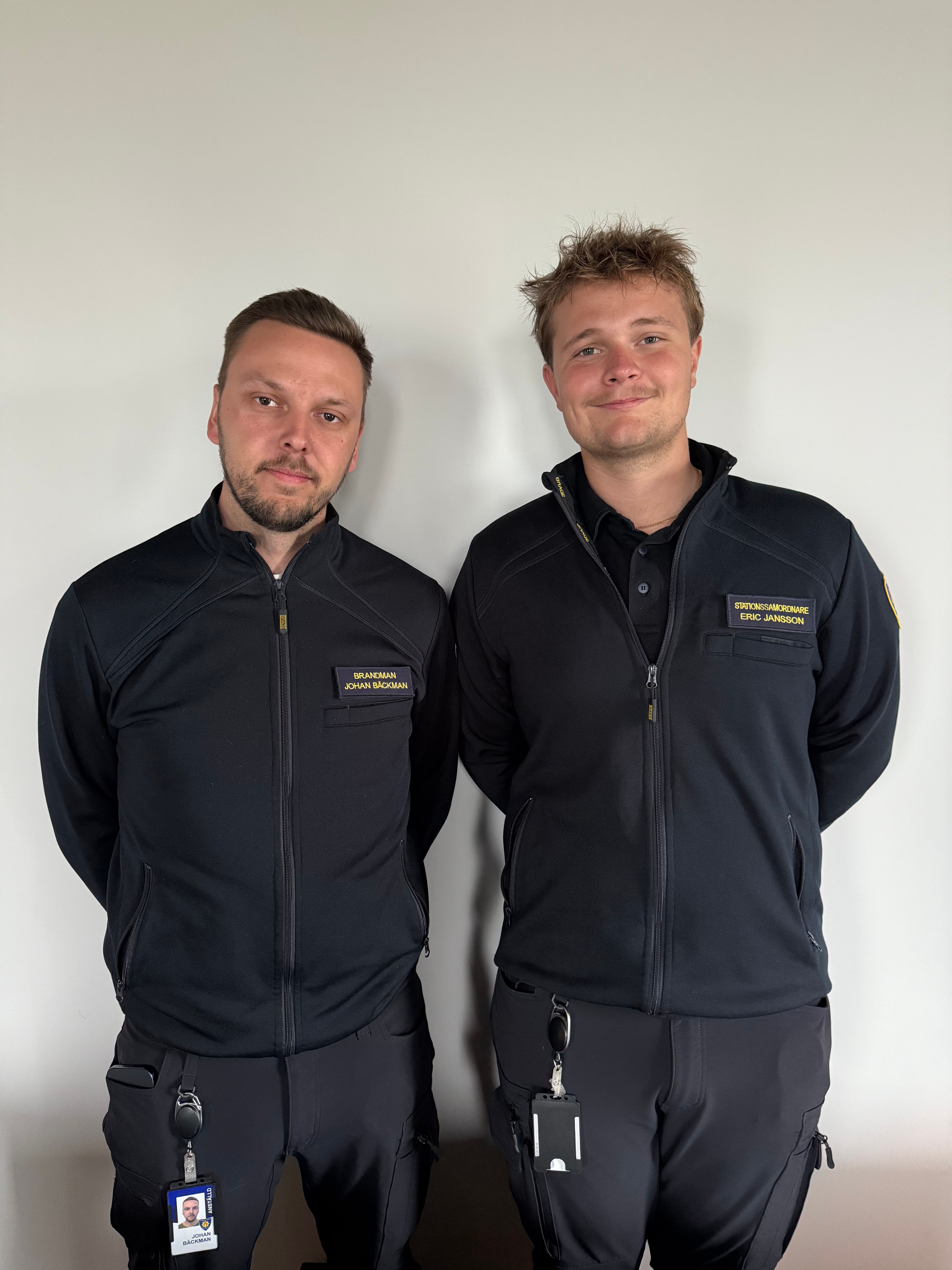 Two men in black uniform jackets with name tags stand side-by-side against a white wall.