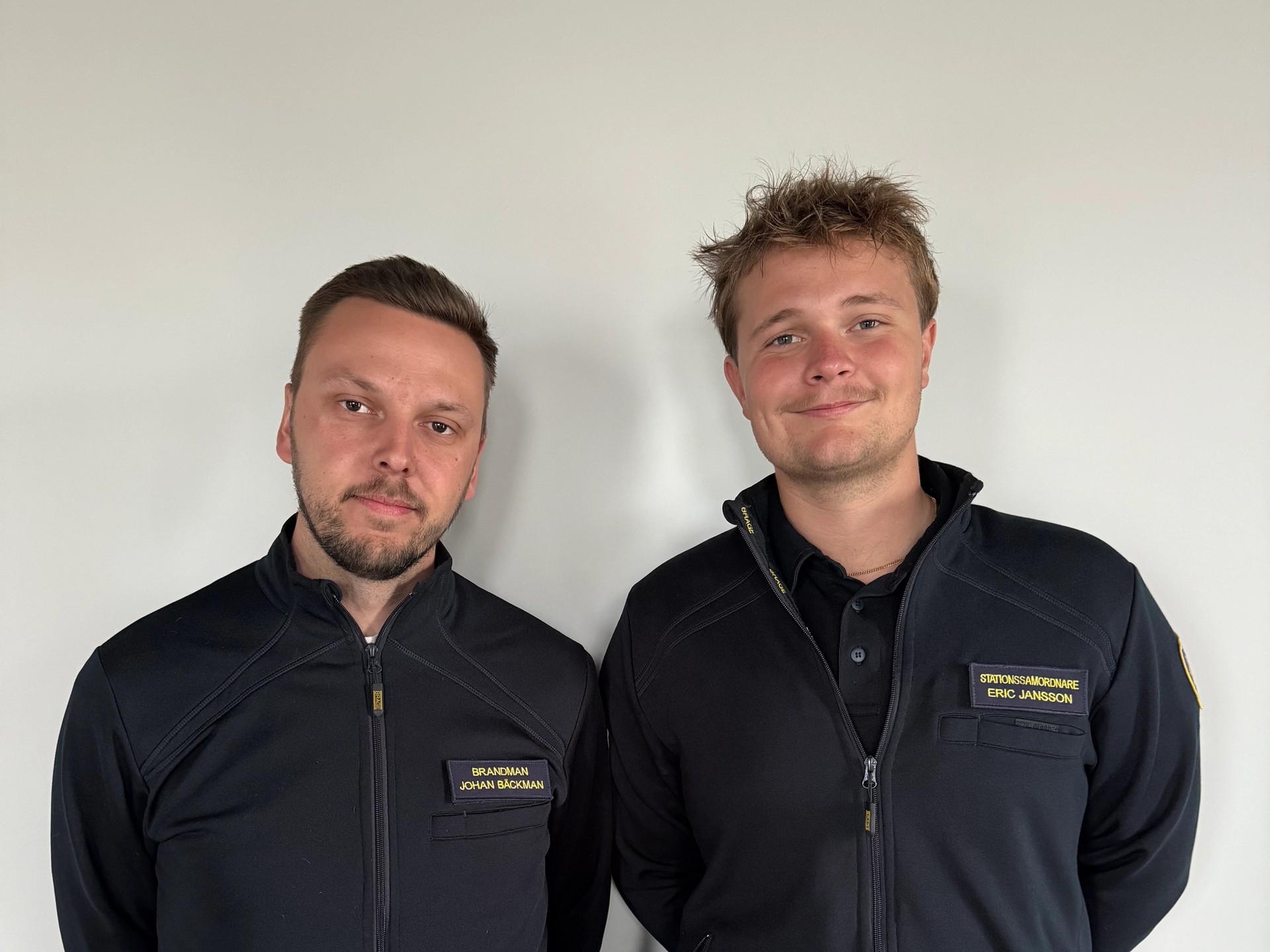 Two men in black uniform jackets with name tags stand side-by-side against a white wall.