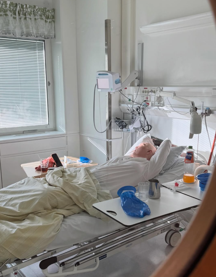 En person ligger i en sjukhussäng, omgiven av medicinsk utrustning och sängbord med drycker.