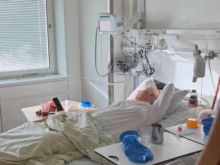 En person ligger i en sjukhussäng, omgiven av medicinsk utrustning och sängbord med drycker.