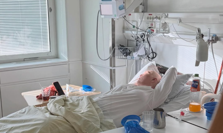 En person ligger i en sjukhussäng med händerna bakom huvudet, med medicinsk utrustning synlig i närheten.