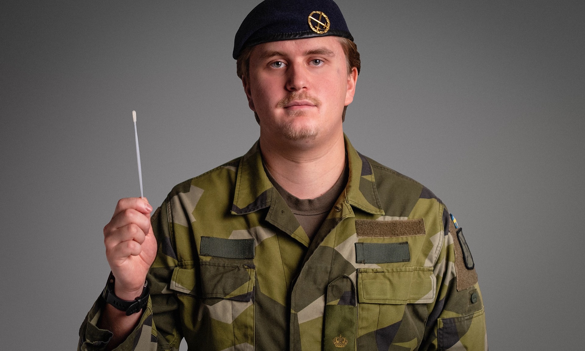 En man i militäruniform och basker håller upp en näsprovpinne.